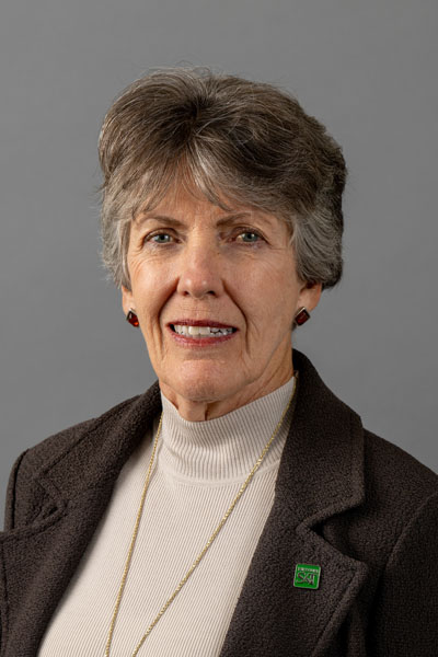 Portrait of a woman with short brown hair, dark brown blazer, tan turtleneck shirt, and a long gold necklace.